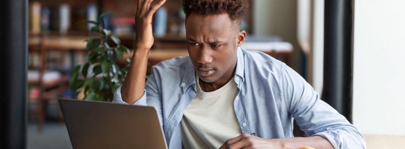 Irritated African American guy using laptop to work on difficult business project at urban cafe