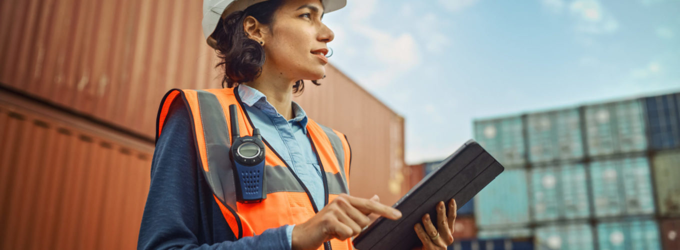 Smiling Portrait of a Latin Female Industrial Engineer in White Hard Hat, High-Visibility Vest Working on Tablet Computer. Inspector or Safety Supervisor in Container Terminal.