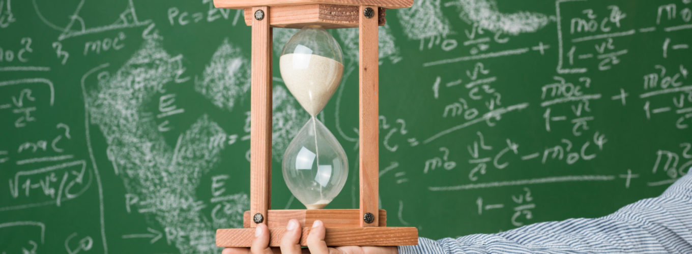 Photo of hour glass in human hand in front of green chalkboard with written mixed mathematical formulas. Einstein's formula E=mc2 is written in big letters. The blackboard is full of formulas. No copy space is seen in frame. Shot with a medium format camera.